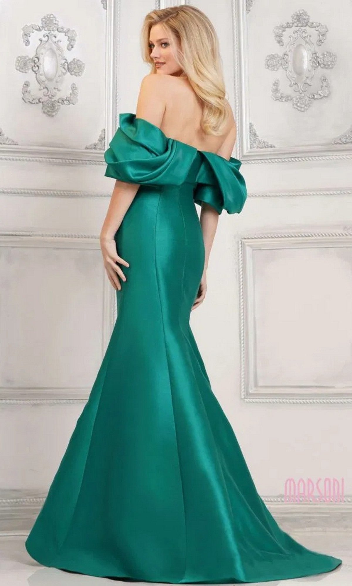 A woman in the Marsoni MV1290 long formal dress, featuring an off-the-shoulder, floor-length green satin mermaid silhouette, stands in an elegant room with ornate white wall panels.